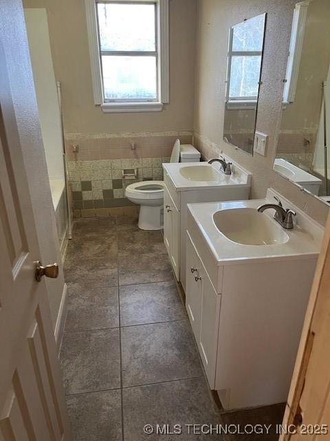 full bath featuring tile walls, a bath, toilet, vanity, and wainscoting