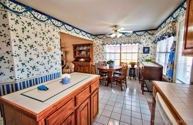 kitchen with wallpapered walls, brown cabinetry, tile counters, light tile patterned flooring, and a ceiling fan