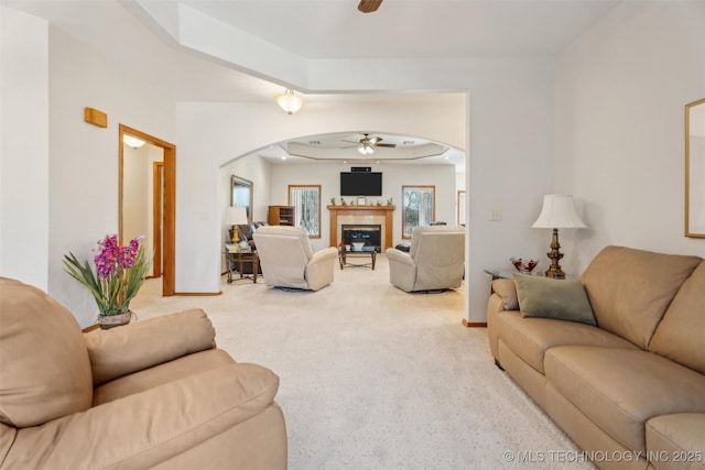 living area with a glass covered fireplace, arched walkways, baseboards, light colored carpet, and ceiling fan