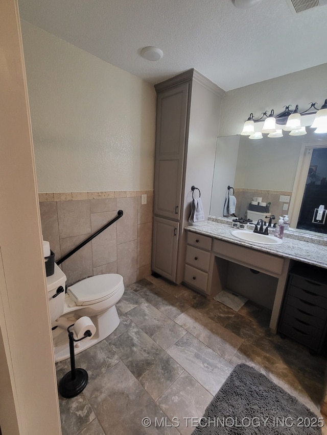 bathroom featuring vanity, a wainscoted wall, a textured ceiling, tile walls, and toilet