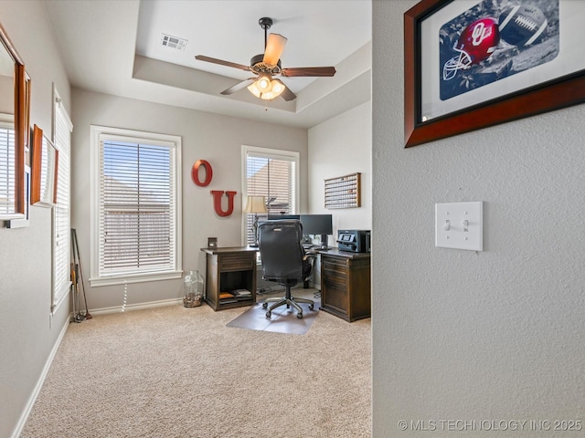 carpeted office space featuring visible vents, a raised ceiling, baseboards, and a ceiling fan