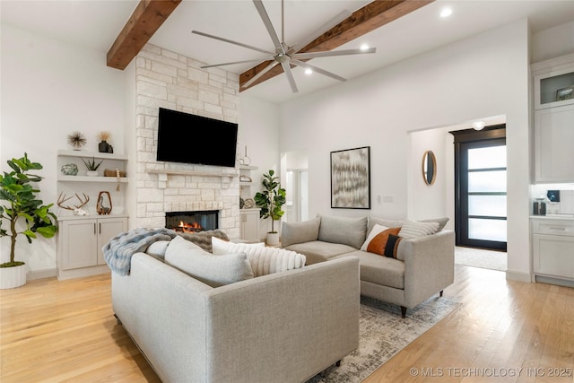 living area with beamed ceiling, a stone fireplace, light wood-style flooring, and ceiling fan