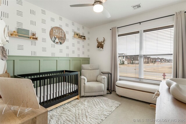 carpeted bedroom with visible vents, wallpapered walls, ceiling fan, a nursery area, and wainscoting