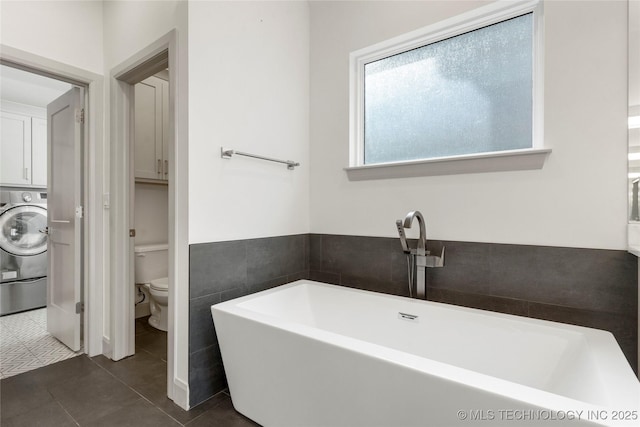 full bath featuring tile patterned flooring, a freestanding bath, toilet, washer / dryer, and tile walls