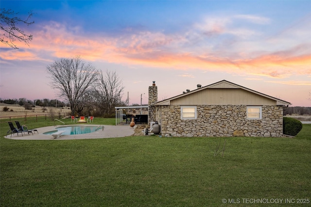 back of property at dusk with stone siding, a lawn, a chimney, and a patio area