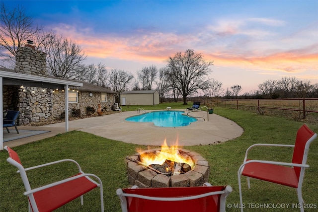 pool at dusk with a patio, fence, an outdoor fire pit, a yard, and an outdoor structure