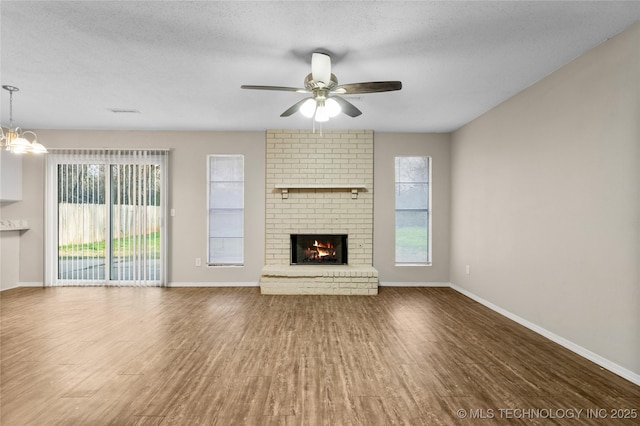 unfurnished living room with a brick fireplace, baseboards, ceiling fan with notable chandelier, wood finished floors, and a textured ceiling