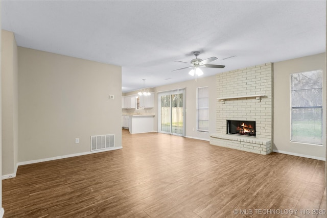 unfurnished living room with visible vents, ceiling fan with notable chandelier, wood finished floors, baseboards, and a brick fireplace