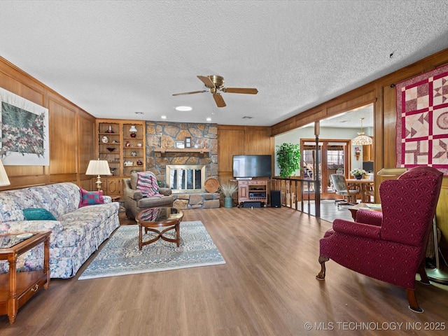 living area with wood walls, a stone fireplace, wood finished floors, a textured ceiling, and a ceiling fan