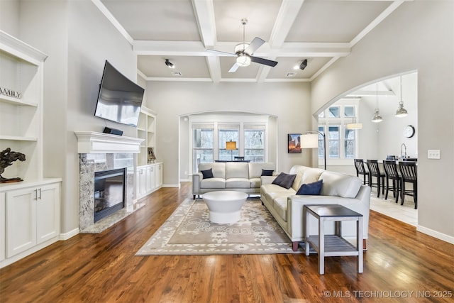 living area featuring wood finished floors, a ceiling fan, beam ceiling, arched walkways, and a high end fireplace