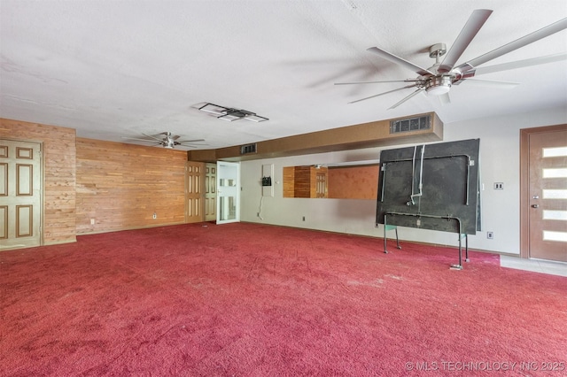 unfurnished living room featuring visible vents, carpet floors, wooden walls, and a ceiling fan