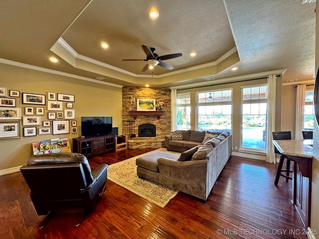 living room with a stone fireplace, ceiling fan, a tray ceiling, and dark wood-style flooring