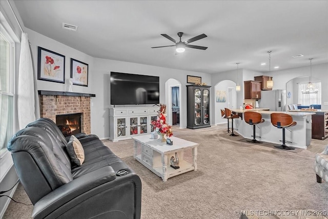 living area with ceiling fan, arched walkways, light carpet, a brick fireplace, and visible vents