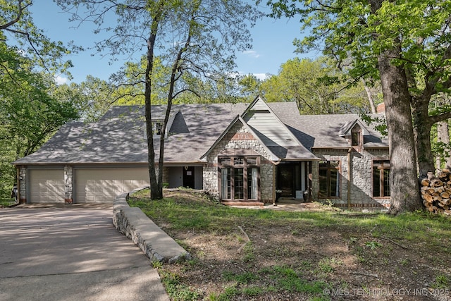 tudor-style house featuring stone siding, an attached garage, a shingled roof, and driveway
