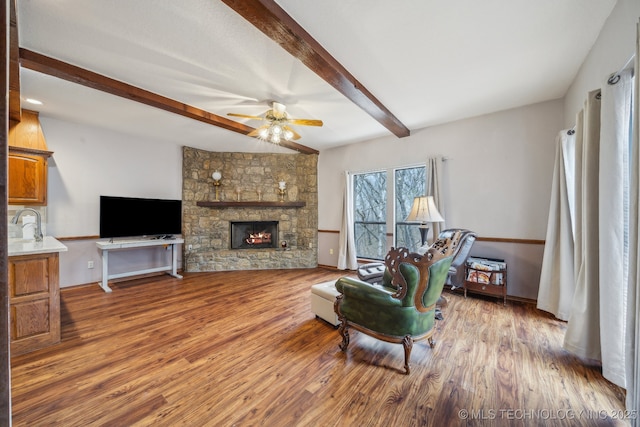 living area featuring wood finished floors, a stone fireplace, beamed ceiling, baseboards, and ceiling fan