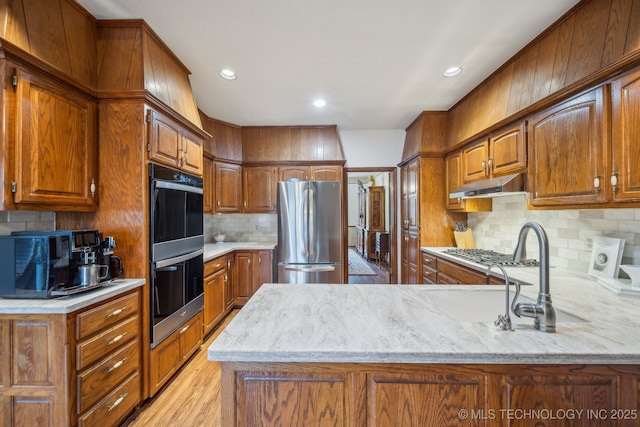 kitchen with under cabinet range hood, decorative backsplash, a peninsula, brown cabinetry, and appliances with stainless steel finishes