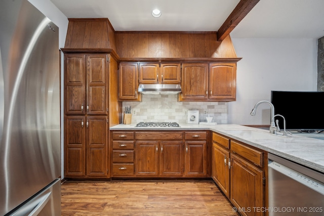 kitchen featuring stainless steel appliances, a sink, light wood-type flooring, backsplash, and under cabinet range hood