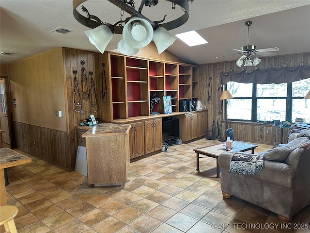 living room featuring a wainscoted wall, visible vents, wooden walls, ceiling fan, and vaulted ceiling with skylight