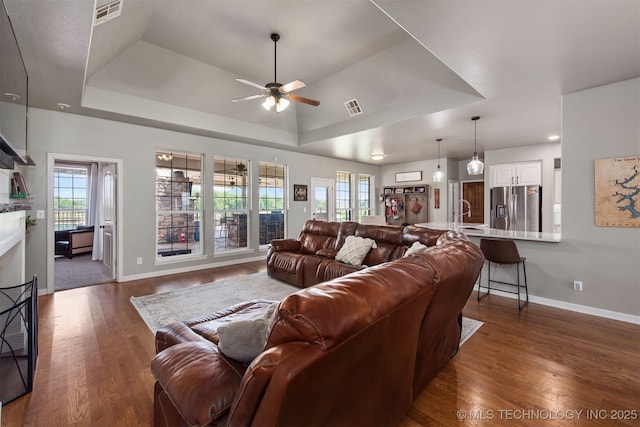 living room featuring ceiling fan, dark wood-style flooring, visible vents, a raised ceiling, and baseboards