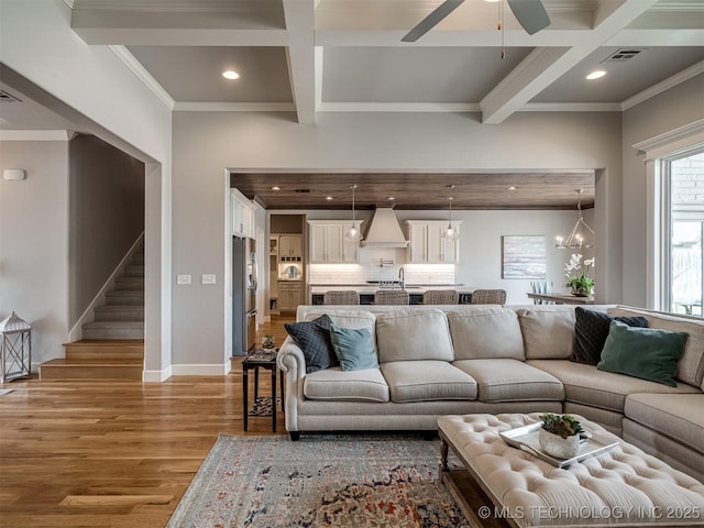 living room with coffered ceiling, stairway, light wood-type flooring, ceiling fan with notable chandelier, and beamed ceiling