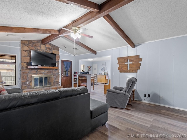 living area with lofted ceiling with beams, a textured ceiling, wood finished floors, a ceiling fan, and a stone fireplace