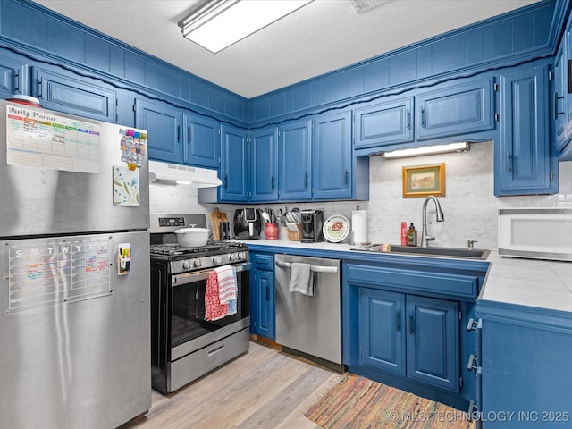 kitchen featuring light countertops, a sink, under cabinet range hood, stainless steel appliances, and blue cabinetry