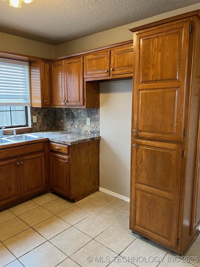 kitchen with light tile patterned flooring, a sink, tasteful backsplash, brown cabinetry, and dark countertops