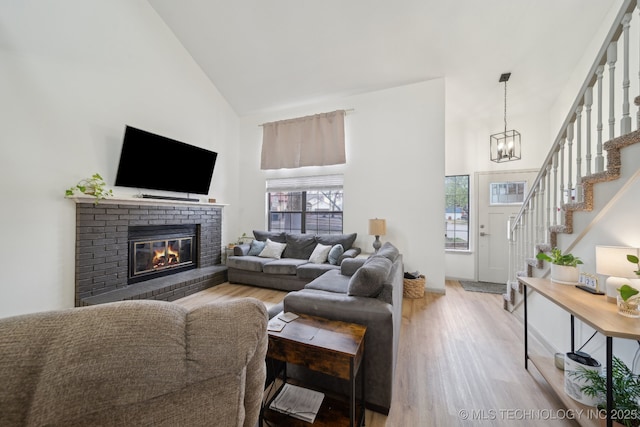 living room with a brick fireplace, high vaulted ceiling, a chandelier, stairs, and light wood-type flooring