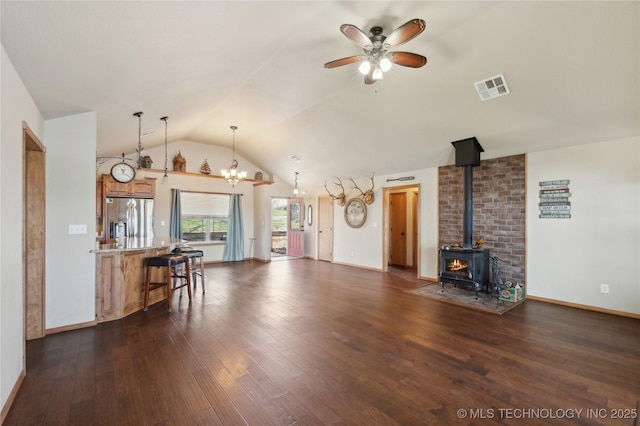 unfurnished living room featuring vaulted ceiling, a wood stove, ceiling fan with notable chandelier, visible vents, and dark wood-type flooring