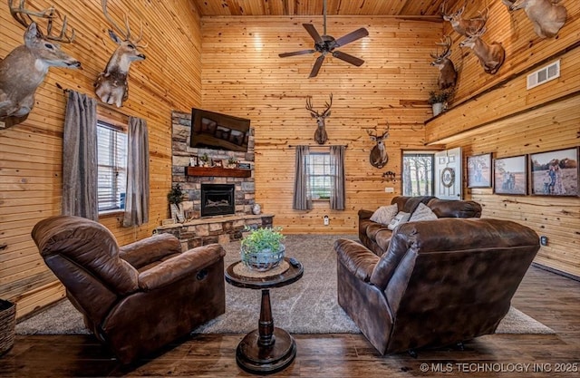 living area with wooden walls, visible vents, a ceiling fan, a stone fireplace, and wood finished floors