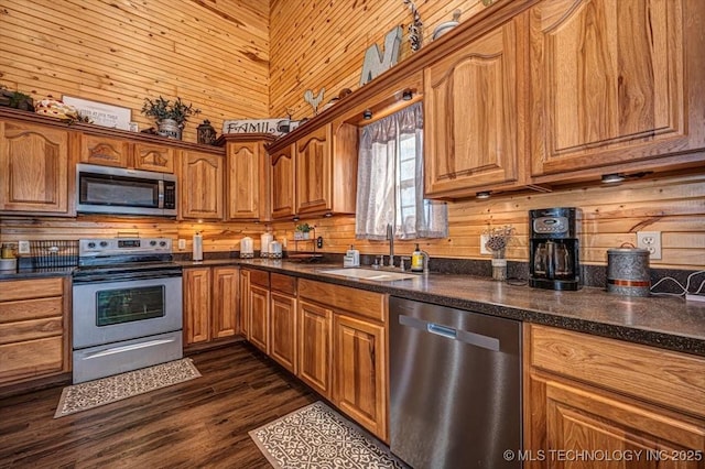 kitchen featuring a sink, wood walls, appliances with stainless steel finishes, brown cabinets, and dark wood-style flooring