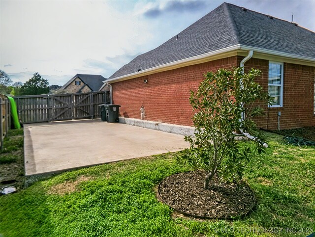 exterior space with a gate, fence, brick siding, a shingled roof, and a patio