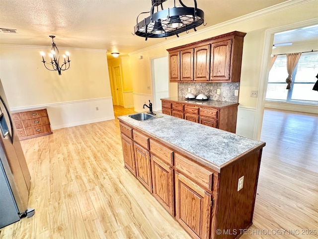 kitchen featuring a notable chandelier, a sink, a kitchen island with sink, crown molding, and light wood-style flooring