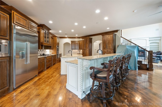 kitchen with built in appliances, arched walkways, decorative backsplash, wood finished floors, and a center island with sink