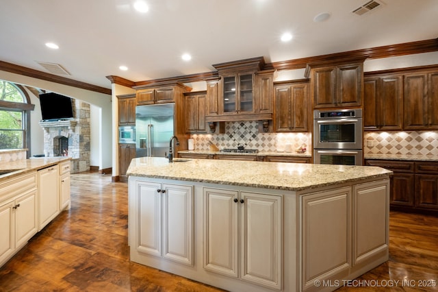 9931 N Chadford Rd, Owasso, OK 74055 - kitchen featuring built in appliances, a kitchen island with sink, ornamental molding, glass insert cabinets, and decorative backsplash