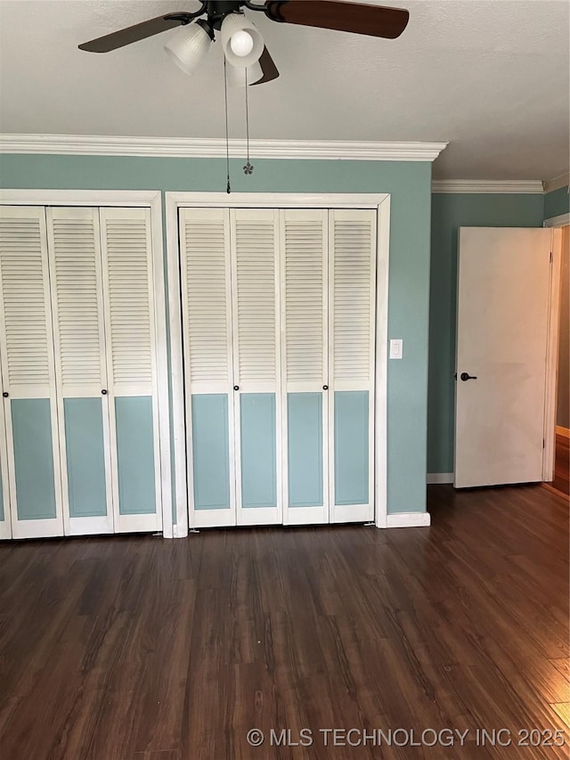 unfurnished bedroom featuring crown molding, two closets, ceiling fan, baseboards, and dark wood-style flooring