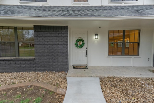 view of exterior entry featuring a porch, roof with shingles, and brick siding