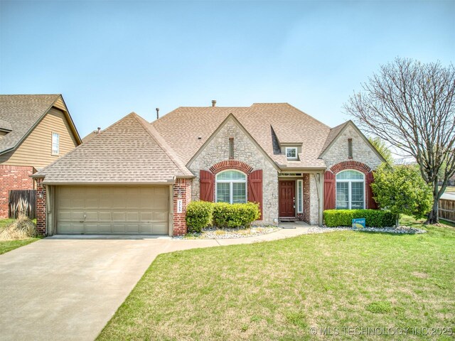 french country home featuring a shingled roof, a front yard, stone siding, an attached garage, and concrete driveway