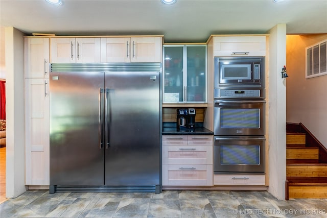 kitchen with built in appliances, glass insert cabinets, visible vents, and dark countertops