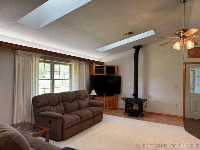 living area with vaulted ceiling with skylight, a wood stove, ceiling fan, visible vents, and light tile patterned flooring