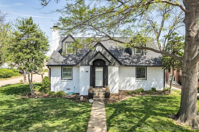 tudor house featuring a front yard, brick siding, a chimney, and a shingled roof