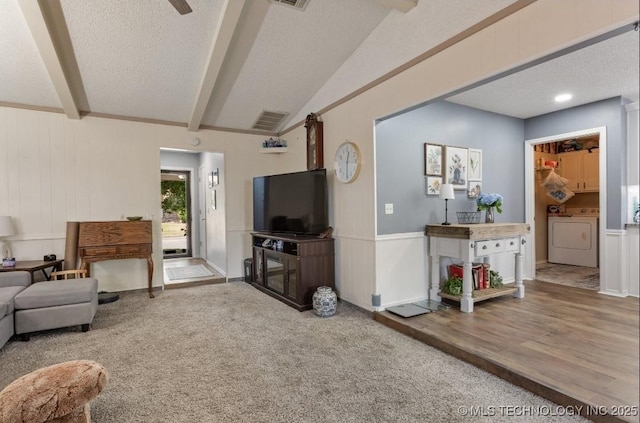 carpeted living area featuring a decorative wall, a textured ceiling, vaulted ceiling with beams, washing machine and dryer, and visible vents