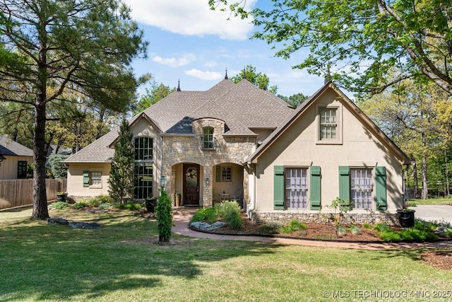 french country style house featuring a front lawn, stone siding, roof with shingles, stucco siding, and fence