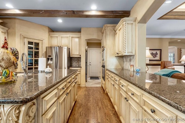 kitchen with beamed ceiling, cream cabinetry, backsplash, arched walkways, and appliances with stainless steel finishes