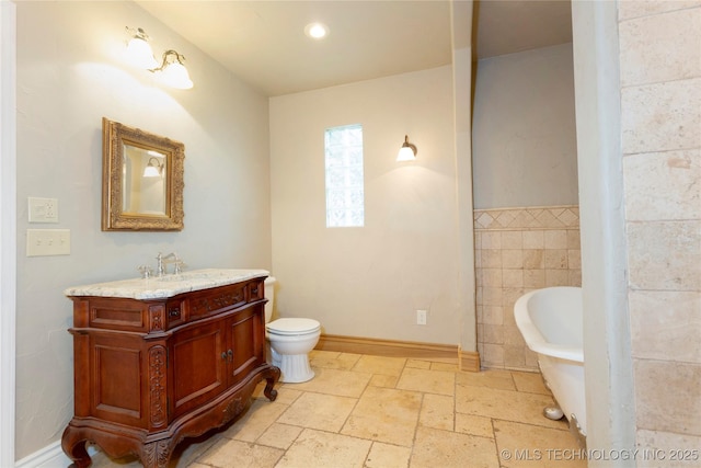 bathroom featuring toilet, vanity, stone tile flooring, baseboards, and a soaking tub