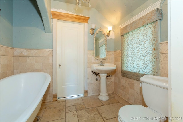 full bathroom with a freestanding bath, toilet, tile walls, wainscoting, and stone tile floors