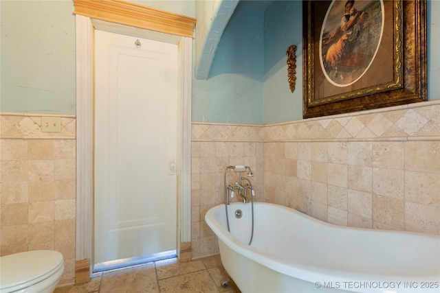 bathroom with tile walls, a wainscoted wall, a freestanding tub, and toilet