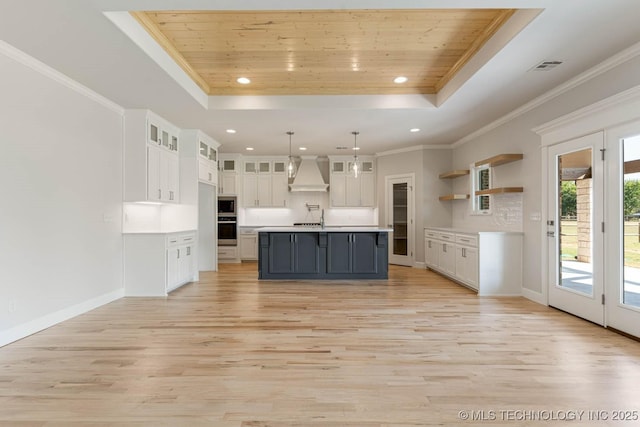 kitchen featuring stainless steel oven, a tray ceiling, light wood-style floors, and premium range hood