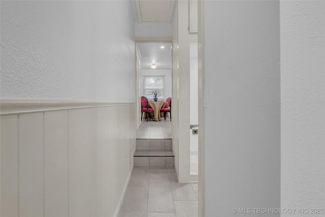 corridor with attic access, a wainscoted wall, a textured wall, and light tile patterned flooring