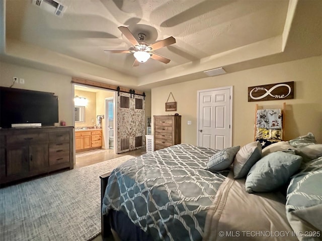 bedroom featuring a barn door, a tray ceiling, visible vents, a ceiling fan, and ensuite bathroom